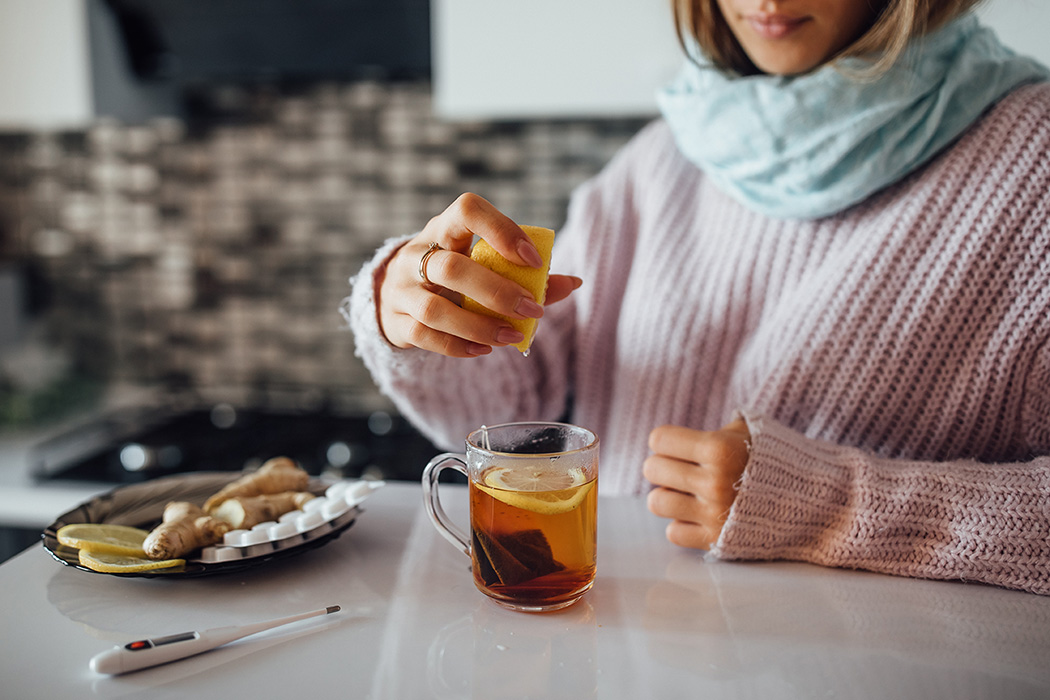 Prävention mit Profil: Was Marken von Erkältungstypen lernen können Eine Frau in warmem Pullover und Schal bereitet eine Tasse Tee mit Zitrone zu; auf dem Tisch liegen Ingwer, Zitronenscheiben, Tabletten und ein Thermometer.