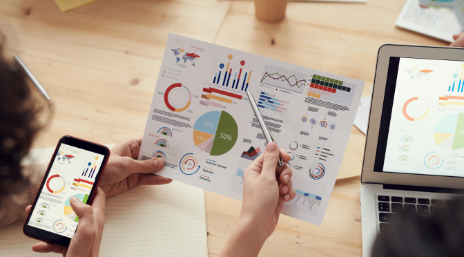 Brandtracking: Person hält ein Papier mit bunten Diagrammen, neben Handy und Laptop, die auch Diagramme zeigen. 