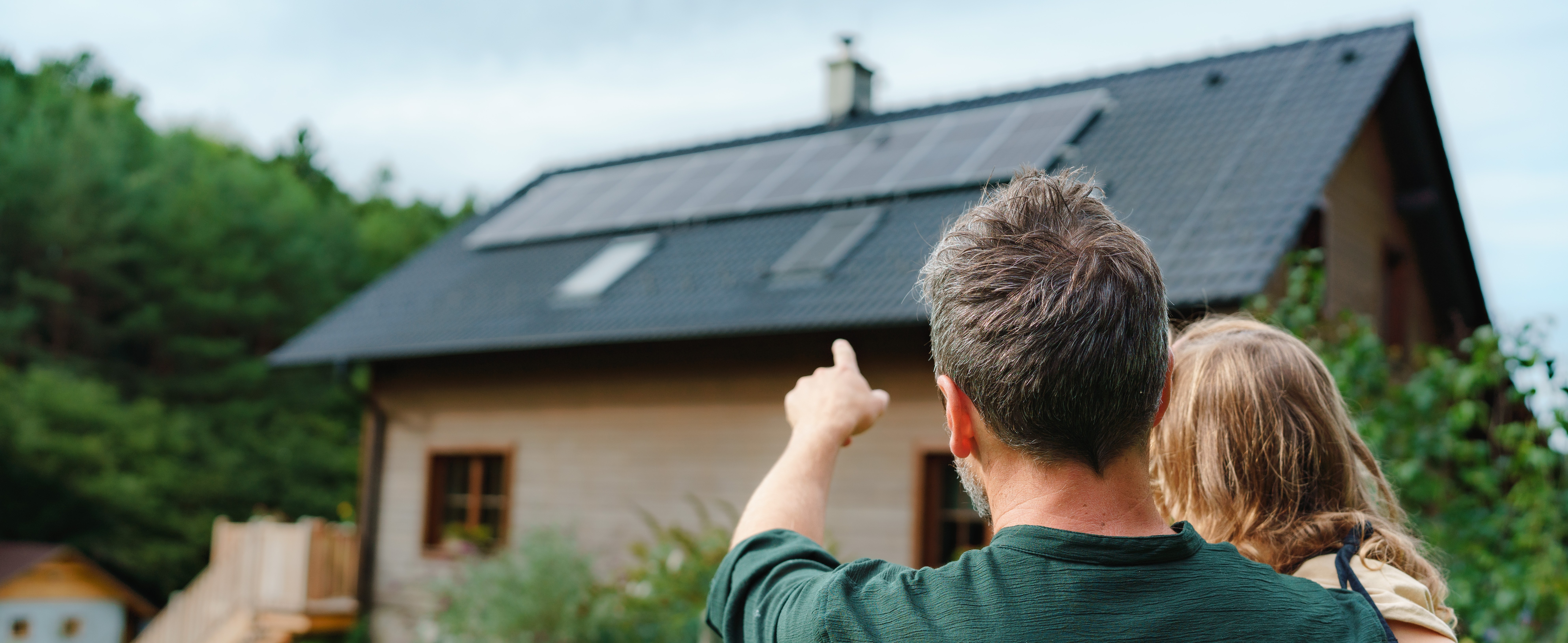 Zwei Personen von hinten, die auf ein Haus mit Solarpanels auf dem Dach blicken; eine Person deutet mit dem Finger auf die Paneele