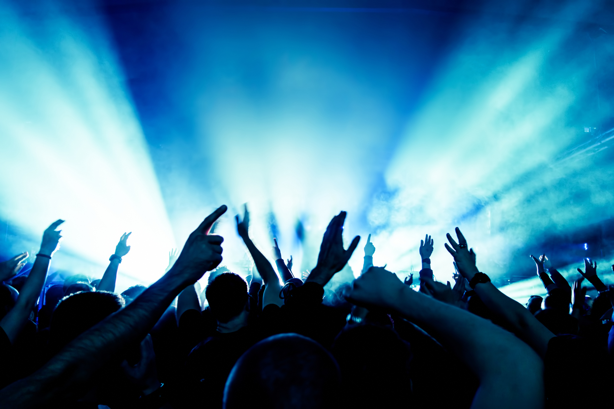 Menschenmenge bei einem Konzert mit erhobenen Händen, im Hintergrund helle Bühnenlichter in Blau und Weiß.