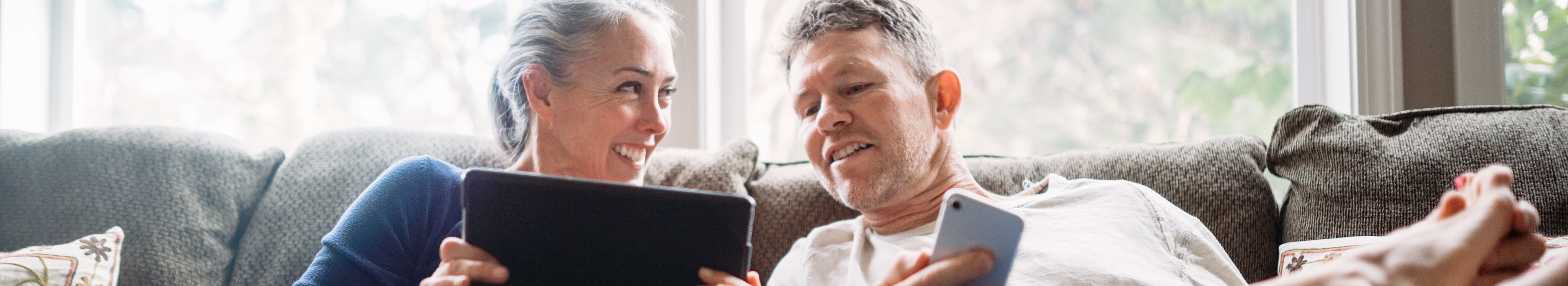 Zwei Personen, die auf einem Sofa sitzen und die Frau einem Mann etwas auf einem Tablet zeigt, während er ein Smartphone in der Hand hält.