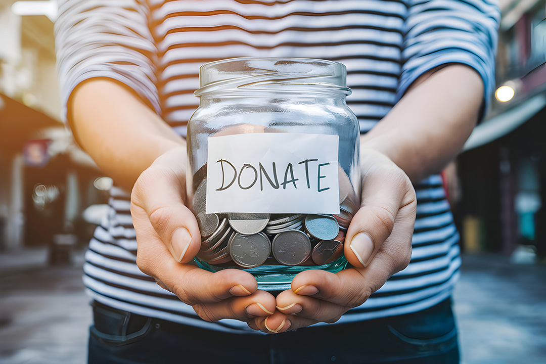Zwei Hände, die ein Glas mit Münzen und einem Zettel mit der Aufschrift „DONATE“ nach vorne halten.