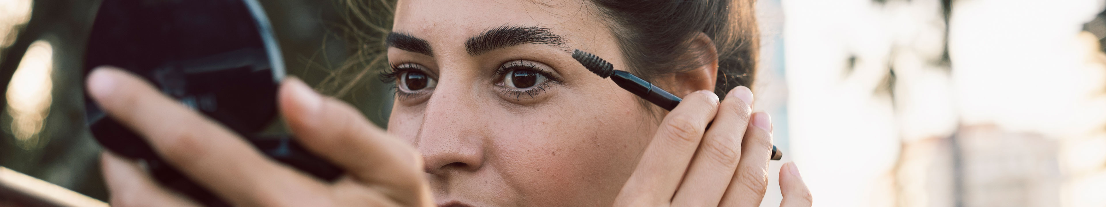 Eine Frau schaut in einen Kosmetik-Handspiegel und nutzt ein Beauty-Produkt.