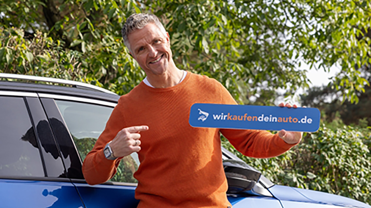 Ein Mann steht vor einem Auto und zeigt auf ein Schild welches er in der anderen Hand hält.