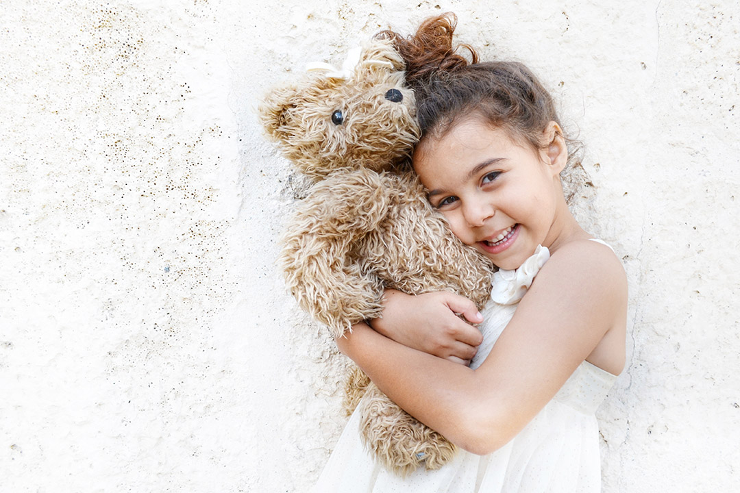 Kleines Mädchen in einem weißen Kleid, das liebevoll einen flauschigen Teddybär umarmt.