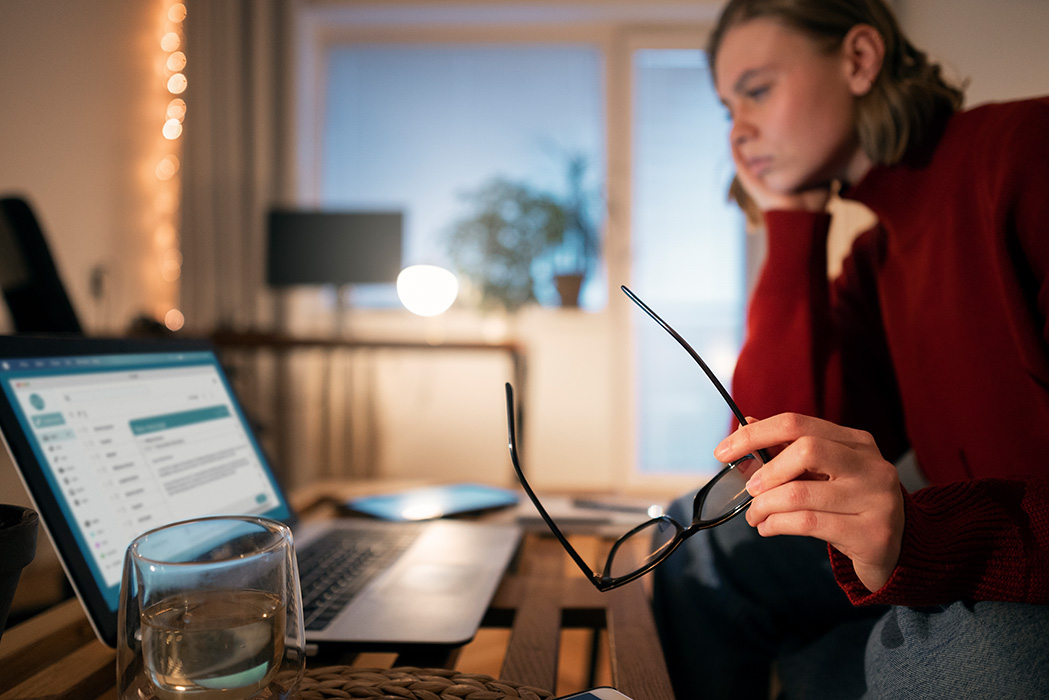 Frau sitzt an einem Tisch vor einem geöffneten Laptop und hält eine Brille in der Hand.