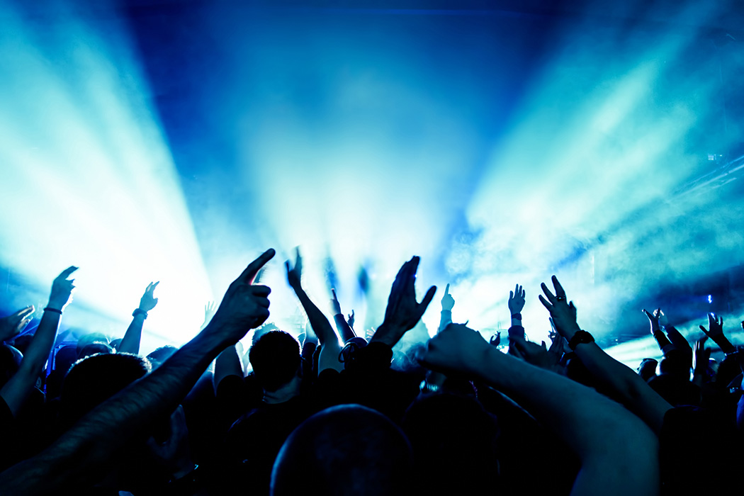 Menschenmenge bei einem Konzert mit erhobenen Händen, im Hintergrund helle Bühnenlichter in Blau und Weiß.
