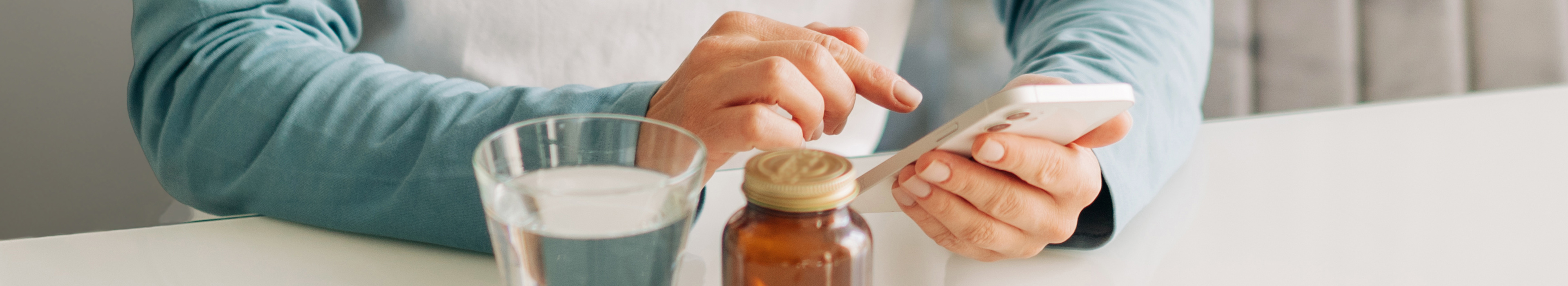 Eine Hand, die ein Smartphone hält, neben einem Glas Wasser und einer Medikamentenflasche.