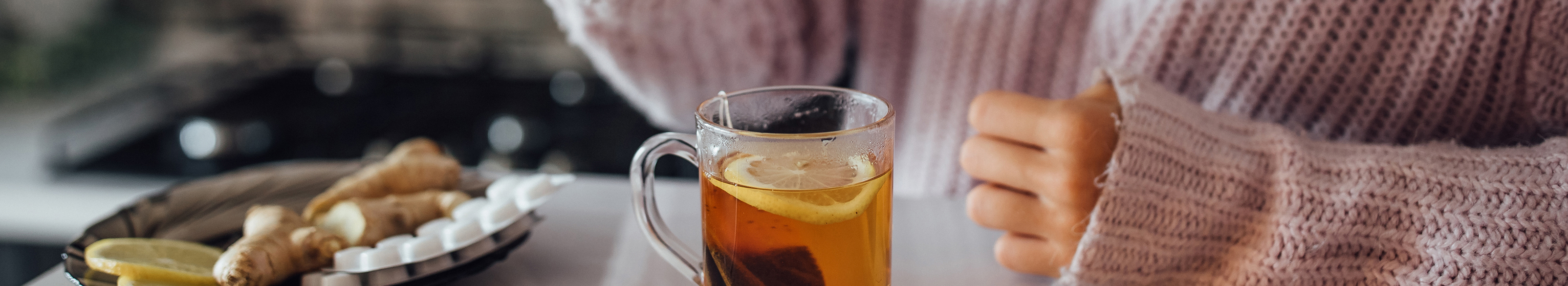 Eine Hand in einem gestrickten Ärmel, die neben einer Glas-Tasse mit heißem Tee und Zitronenscheibe liegt; daneben frischer Ingwer und Tabletten auf einem Teller.