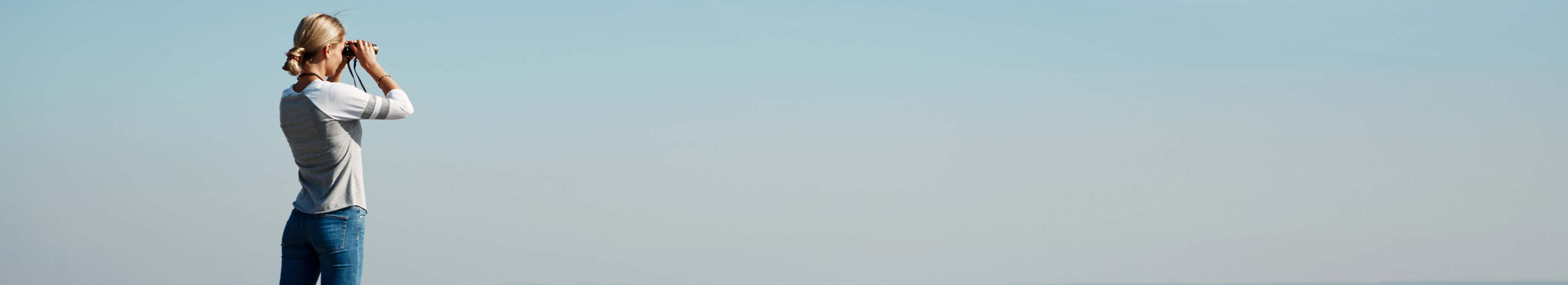 Person, die durch ein Fernglas blickt vor einem blauen Horizont.